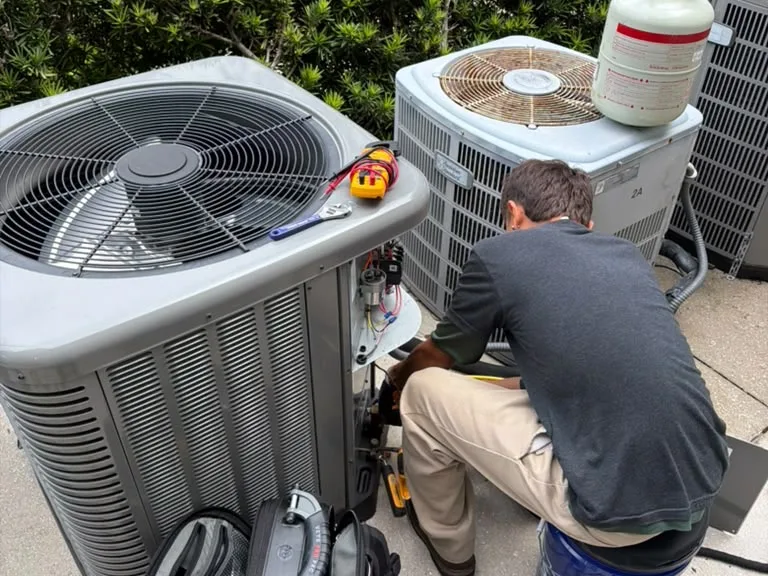 Technician crouched at an outdoor AC condenser beside a hedge during a repair