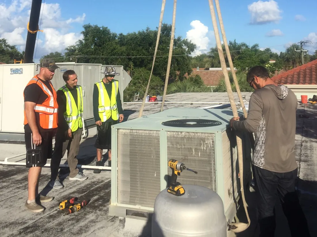 Crew in safety vests on a flat roof beside a packaged rooftop unit on lifting straps