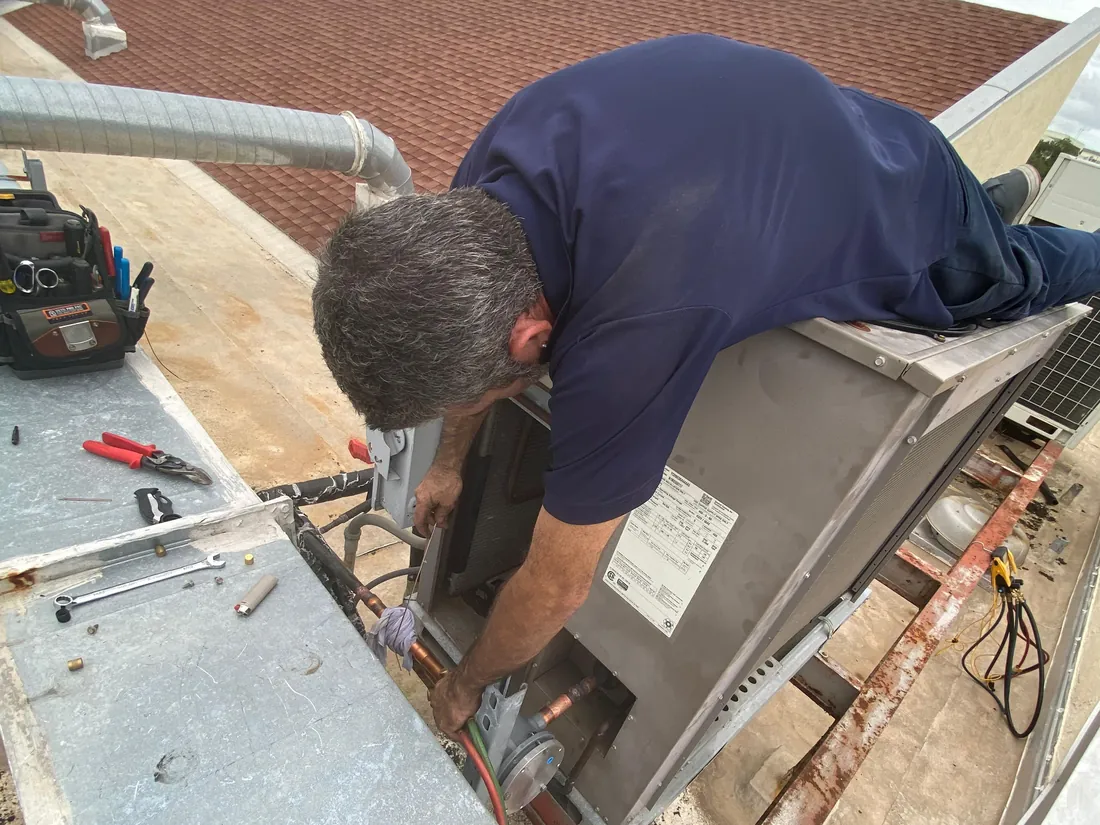 ACR technician leaning over a rooftop unit to work on its copper refrigerant lines, with tools laid out on the ledge beside it