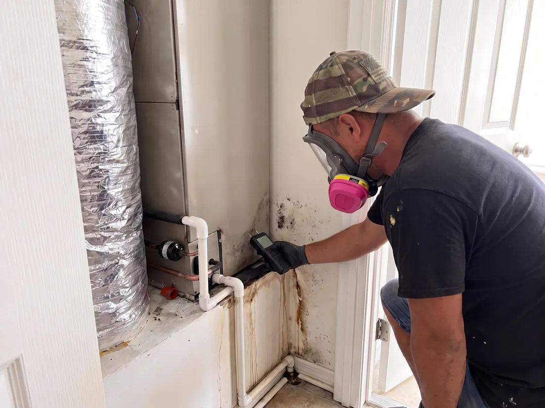 Technician holding a moisture meter to a water-stained, moldy wall beside an air handler and its condensate drain line