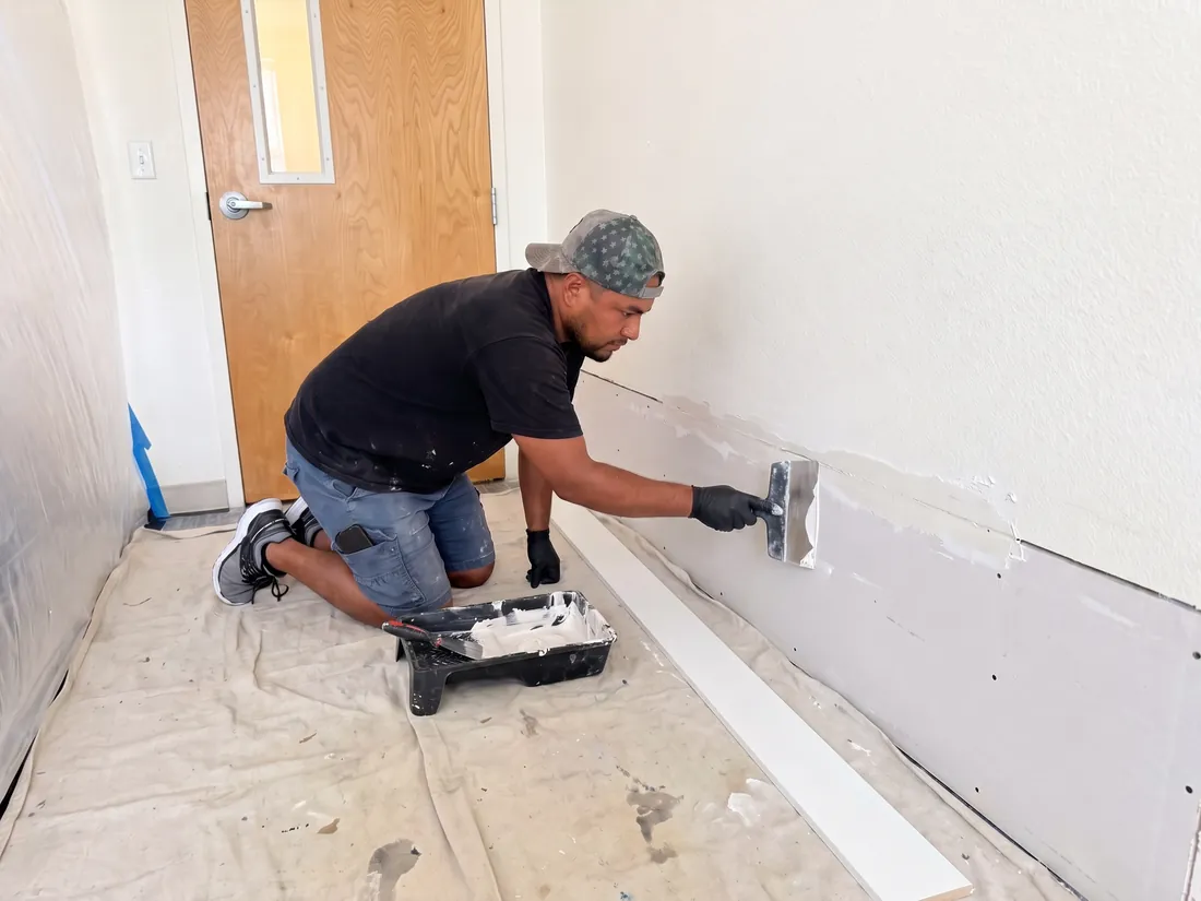 Technician spreading joint compound over new drywall along the bottom of a hallway wall, with a new baseboard on the drop cloth