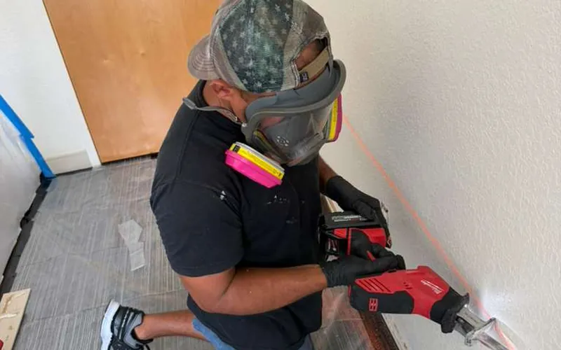 Remediation tech in a full-face respirator cutting out damaged drywall inside a contained work area
