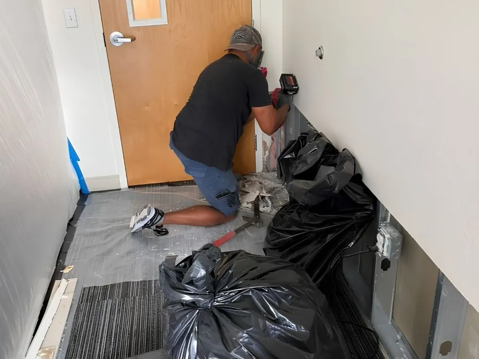 ACR technician removing drywall in a hallway lined with plastic sheeting, with bags of removed material beside him