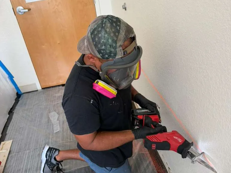 ACR technician in a full-face respirator and gloves cutting along a laser line on a wall, over plastic floor sheeting