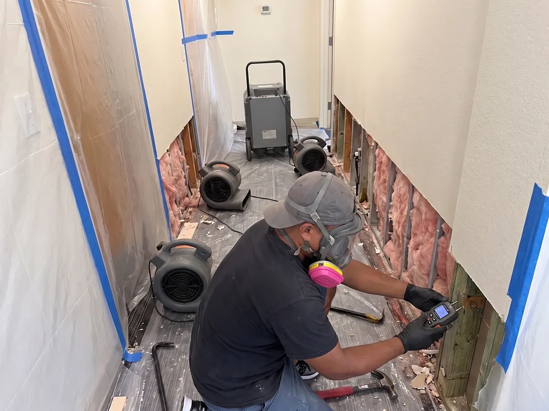Technician checking a wall stud with a moisture meter, with air movers and a dehumidifier in the hallway