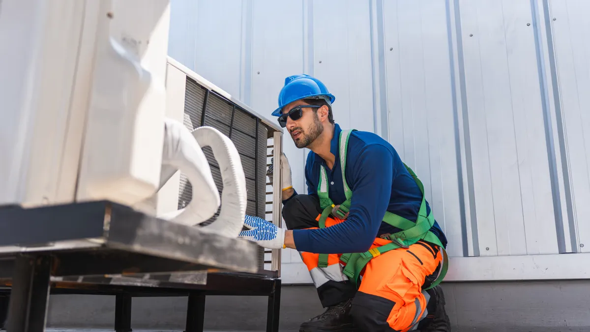 Technician in a hard hat and safety harness kneeling beside a rooftop AC unit