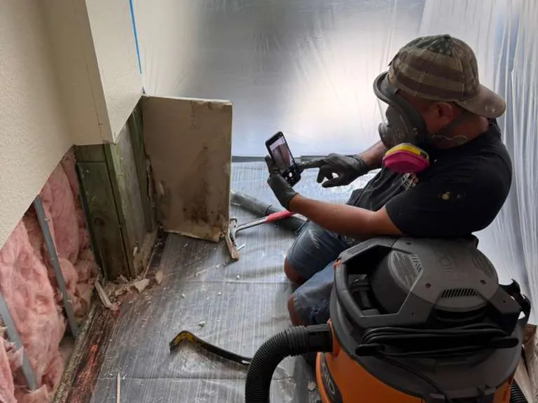 Technician photographing a wall opened to the studs and insulation, with a shop vacuum beside him